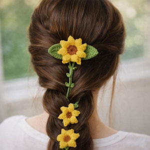 May include: A crocheted sunflower hair accessory, with yellow petals and a brown centre, is woven into a braid. The accessory includes green leaves and a stem, adding a touch of nature-inspired charm to the hairstyle. The hair is a dark brown colour.
