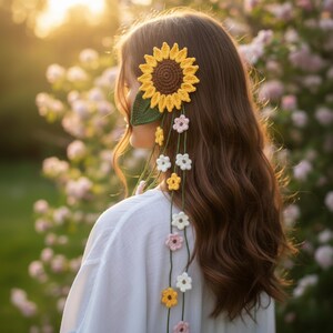 Könnte beinhalten: Gehäkeltes Haaraccessoire mit Sonnenblumen-Design, brauner Mitte und gelben Blütenblättern. Grüne Blätter und Stränge aus gehäkelten Blumen in Weiß, Rosa und Gelb. Das Accessoire wird im Haar getragen, mit einem weichen, verschwommenen floralen Hintergrund.