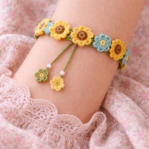 Puede incluir: Pulsera de ganchillo con detalles de flores amarillas y azules. La pulsera está adornada con pequeños colgantes de flores y cuentas blancas. La pulsera se lleva en la muñeca, mostrando el diseño floral hecho a mano.