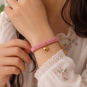 Könnte beinhalten: Ein rosa gewebtes Armband mit einem goldenen Herzanhänger. Das Armband wird mit einer goldenen Kette und einem Verschluss befestigt. Das Armband wird am Handgelenk getragen.