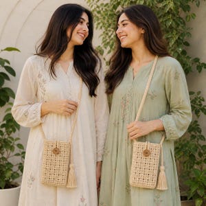 May include: Two women wearing long-sleeve dresses, one cream-colored and the other light green. Each dress has floral embroidery and a woven beige crossbody phone bag with a wooden button and tassel. The bags hang from braided straps.