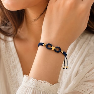 May include: Navy blue macrame bracelet featuring a gold heart charm and gold beads. The bracelet is designed in an infinity symbol shape. The adjustable cord has gold bead accents.