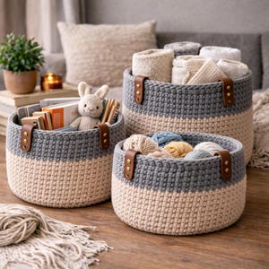 Könnte beinhalten: Drei handgefertigte Häkel-Aufbewahrungskörbe in verschiedenen Größen. Jeder Korb hat ein zweifarbiges Design mit einem grauen oberen Bereich und einem beigen unteren Bereich. Braune Ledergriffe sind mit Messingnieten befestigt. Die Körbe sind mit Garn, Büchern und einem Stoffhasen gefüllt.