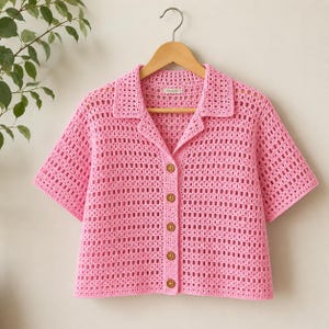May include: A bright pink short-sleeved crochet shirt featuring a classic collar, an open square-stitch pattern, and a front button-down closure with six round, light brown wooden buttons. The garment is displayed on a wooden clothes hanger.