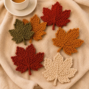 May include: A collection of six crocheted maple leaf coasters in autumn colors including deep red, burnt orange, olive green, and cream. Each leaf features detailed stitched veins and a textured surface, arranged in a cluster on a soft fabric.