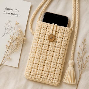 May include: Cream-coloured woven phone crossbody bag with a wooden button closure and a tassel. The bag holds a black smartphone and has a long, braided strap. The text "Enjoy the little things" is on a card.