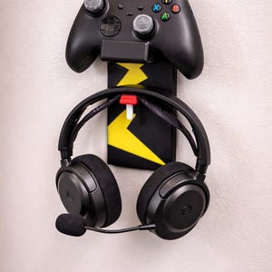 May include: Black gaming controller and headset holder. The holder is black with yellow lightning bolt accents. The controller is black with gray joysticks and colored buttons. The headset is black with a microphone.