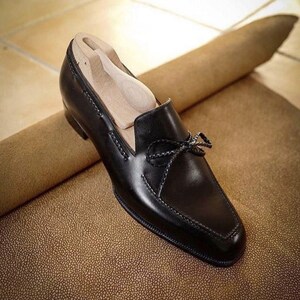 May include: A black leather loafer with a bow detail. The shoe has a square toe and decorative stitching. A wooden shoe tree is inside the shoe. The shoe is on a textured brown surface.