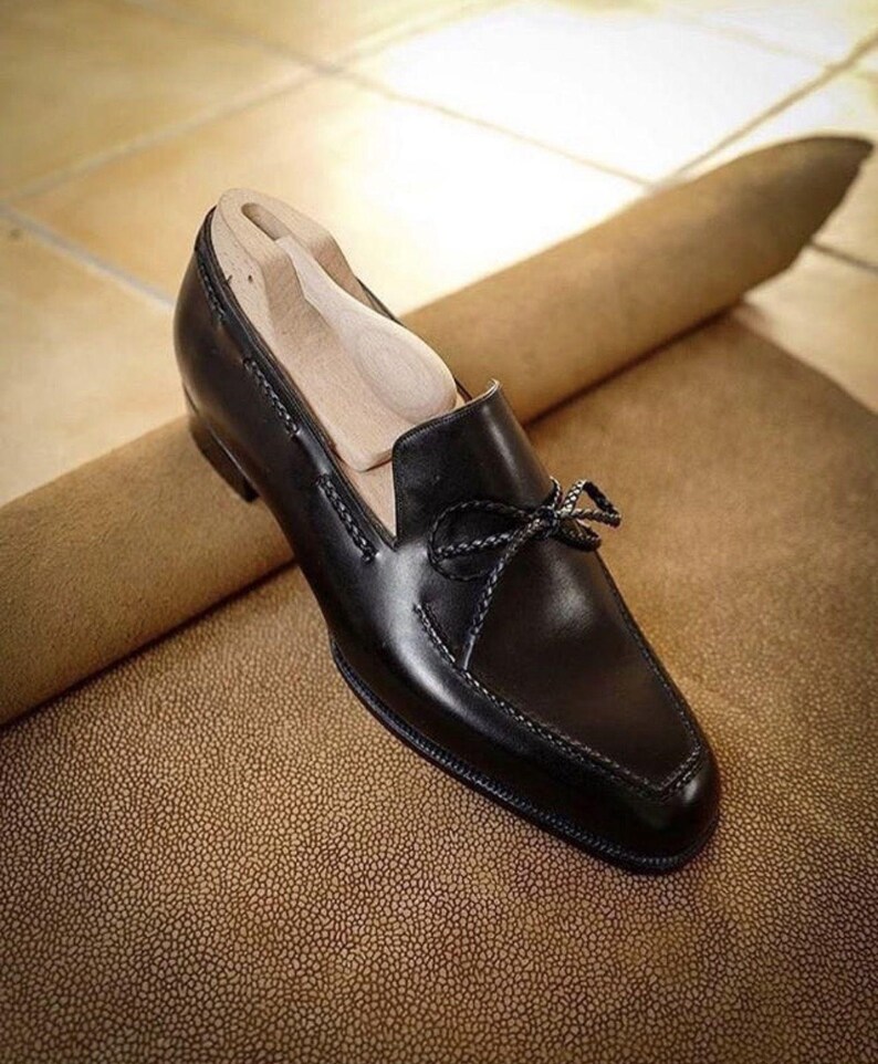 May include: A black leather loafer with a bow detail. The shoe has a square toe and decorative stitching. A wooden shoe tree is inside the shoe. The shoe is on a textured brown surface.