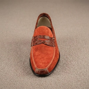 May include: A slip-on loafer shoe featuring a vibrant orange suede upper with a dark brown crocodile-textured leather strap across the vamp. The shoe includes white contrast stitching along the edges and a black sole.