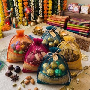 May include: An assortment of colorful gift bags filled with chocolates and seashells. The bags are in shades of orange, pink, teal, and yellow, with gold trim. Various chocolates and nuts are scattered around the bags.
