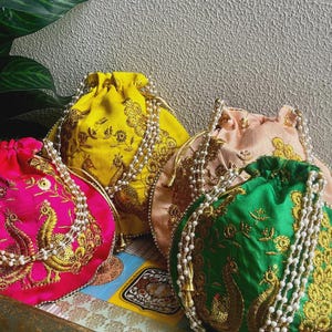 Peut inclure: Quatre sacs à cordon colorés avec des broderies dorées complexes et des perles. Les sacs sont dans des tons de rose, jaune, pêche et vert, chacun avec un motif de paon. Les sacs sont disposés sur un plateau décoratif.