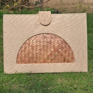 May include: A rectangular beige document holder with a woven, semi-circular design in the center. The holder has a flap closure at the top. The item is made of natural materials and is sitting on green grass.