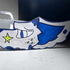 Può includere: Una testa di personaggio dei cartoni animati disegnata a mano, con una base bianca, accenti blu e una stella gialla. Il personaggio ha una faccia sorridente con gli occhi chiusi e piccole stelle gialle intorno al viso. Il design è su sfondo bianco.