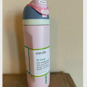 May include: A light pink Owala water bottle with a blue and light pink lid featuring a push button and a lift to unlock latch. The bottle has a white label with green text that reads Hi there. I'm leak proof. I'm insulated. And I have a built-in straw. 24 oz./710ml.