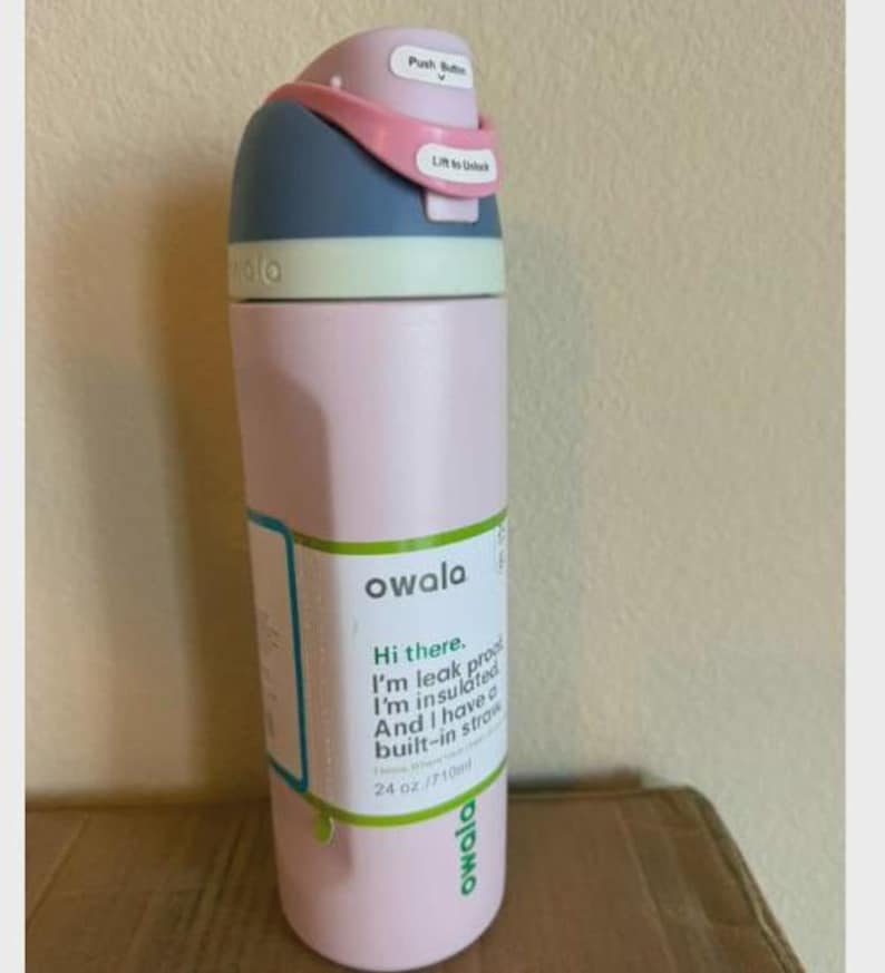 May include: A light pink Owala water bottle with a blue and light pink lid featuring a push button and a lift to unlock latch. The bottle has a white label with green text that reads Hi there. I'm leak proof. I'm insulated. And I have a built-in straw. 24 oz./710ml.