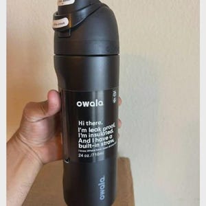 May include: A matte black Owala water bottle with a 24 oz. capacity. The bottle features a black lid with a push button and a lock mechanism. Printed text on the front reads: Hi there. I'm leak proof. I'm insulated. And I have a built-in straw.