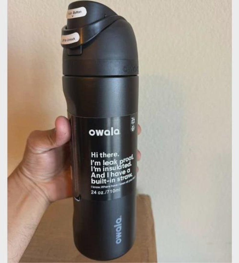 May include: A matte black Owala water bottle with a 24 oz. capacity. The bottle features a black lid with a push button and a lock mechanism. Printed text on the front reads: Hi there. I'm leak proof. I'm insulated. And I have a built-in straw.