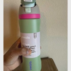 May include: A sage green Owala water bottle with a gray and light green lid featuring a pink accent band. The bottle has a white label with the text Hi there. I'm leak proof. Insulated. Use a straw. and a small 24-hour icon. The brand name Owala is printed in purple.
