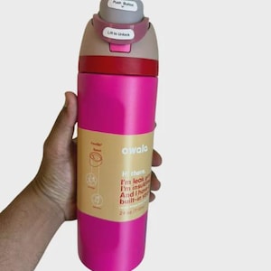 May include: A bright pink Owala water bottle with a 24 oz capacity. The lid features a gray push button and a pink locking mechanism labeled Lift to Unlock. A tan paper label wraps around the center with text stating I'm leak proof, I'm insulated.