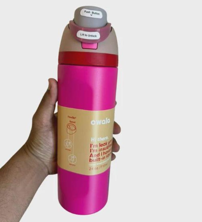 May include: A bright pink Owala water bottle with a 24 oz capacity. The lid features a gray push button and a pink locking mechanism labeled Lift to Unlock. A tan paper label wraps around the center with text stating I'm leak proof, I'm insulated.