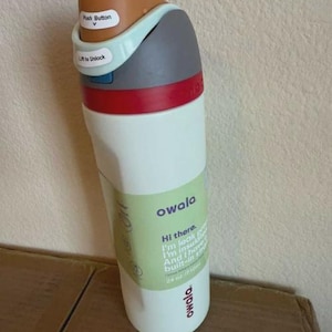 May include: A white Owala water bottle with a 24 oz capacity, featuring a brown push button, a light blue lift-to-unlock latch, a gray cap, and a red accent ring. A light green label on the front displays the brand name and product details.