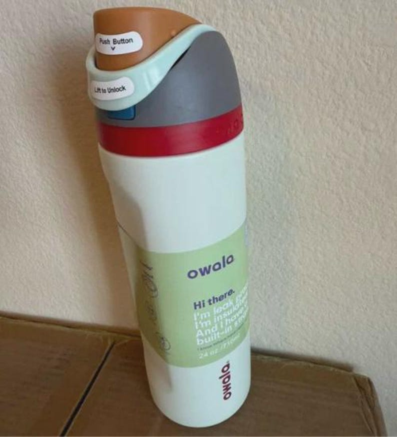 May include: A white Owala water bottle with a 24 oz capacity, featuring a brown push button, a light blue lift-to-unlock latch, a gray cap, and a red accent ring. A light green label on the front displays the brand name and product details.