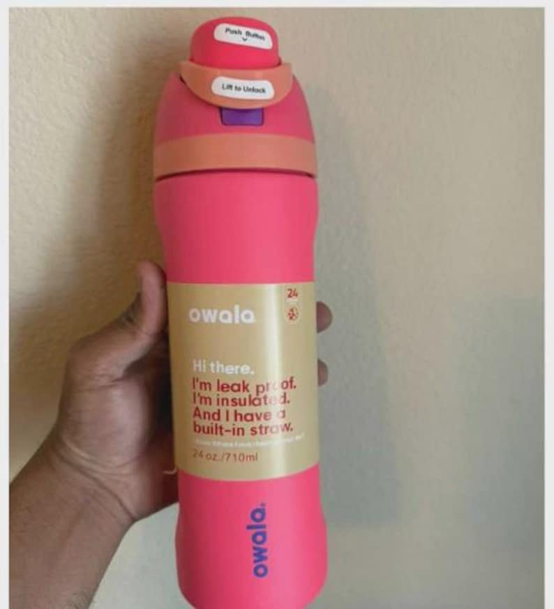 May include: A bright pink Owala water bottle with a peach-colored lid, a purple latch, and a white push button. The bottle features a tan label with the text: Hi there. I'm leak proof. I'm insulated. And I have a built-in straw. 24 oz./710ml.