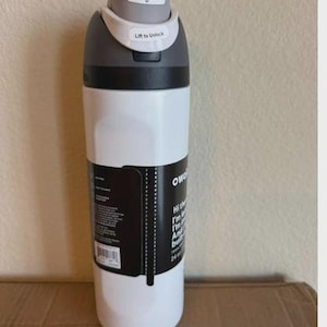 May include: A white Owala water bottle with a gray lid featuring a push button and a lift to unlock mechanism. A black product label is wrapped around the center, displaying white text and a barcode, with a 24 oz capacity clearly printed on it.