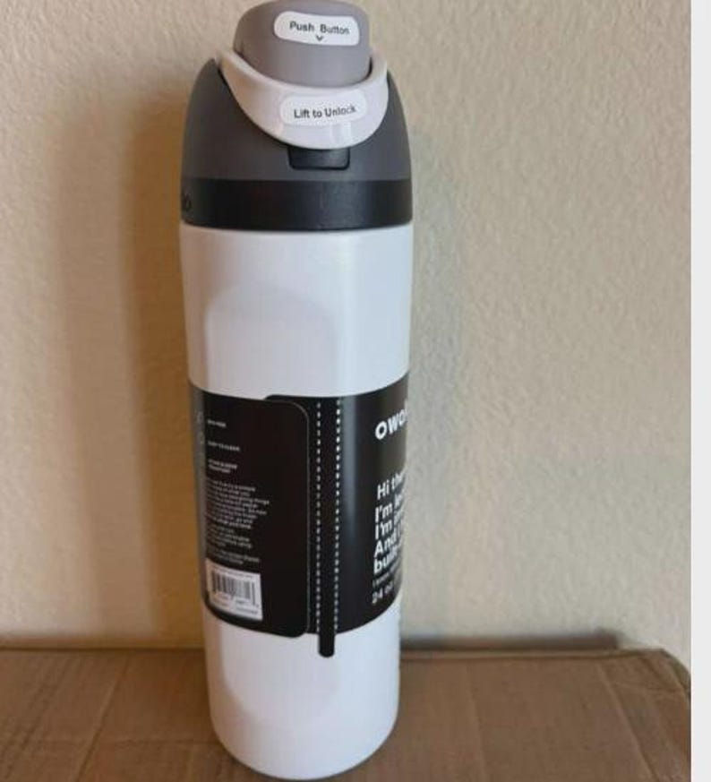 May include: A white Owala water bottle with a gray lid featuring a push button and a lift to unlock mechanism. A black product label is wrapped around the center, displaying white text and a barcode, with a 24 oz capacity clearly printed on it.