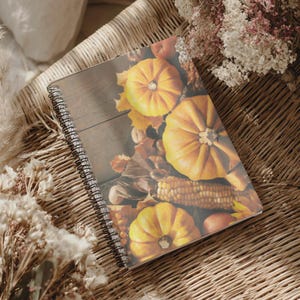Könnte beinhalten: Ein spiralgebundenes Notizbuch mit einem fotografischen Einband, der orangefarbene Kürbisse, getrockneten Mais und Herbstblätter auf einer dunklen Holzoberfläche zeigt. Das Notizbuch hat eine schwarze Metallspirale an der linken Seite.