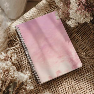 Könnte beinhalten: Ein spiralgebundenes Notizbuch mit einem weichen, rosa-weißen Farbverlauf auf dem Einband, der eine wolkenartige Aquarelltextur aufweist. Das Notizbuch hat eine schwarze Metallspirale an der linken Seite und eine glatte, matte Oberfläche.