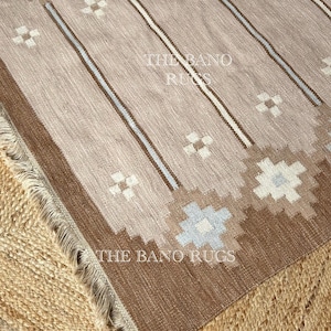 Può includere: Un tappeto intrecciato nei toni del beige, marrone e azzurro con motivi geometrici, strisce verticali e un bordo sfrangiato. Il testo THE BANO RUGS è stampato due volte sulla superficie del tappeto in un carattere bianco senza grazie.
