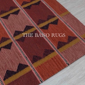 May include: Close-up of a colorful rug with a geometric pattern. The rug features a repeating design of triangles and stripes in shades of red, orange, and brown, with accents of yellow and black. The text "THE BANO RUGS" is visible.