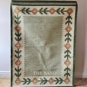 May include: A rectangular area rug featuring a solid sage green centre panel surrounded by a cream border with a repeating pattern of orange flowers and dark green foliage. The text THE BANO RUGS is printed in white on the lower centre of the rug.