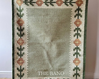 Puede incluir: Una alfombra rectangular con un panel central de color verde salvia sólido rodeado por un borde crema con un patrón repetitivo de flores naranjas y follaje verde oscuro. El texto THE BANO RUGS está impreso en blanco en la parte inferior.
