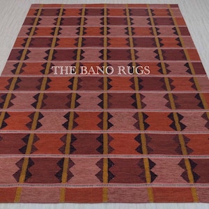 May include: A rectangular area rug featuring a repeating grid pattern in shades of terracotta, deep red, and mustard yellow. The design includes dark geometric diamond-like shapes centered within each block. The text THE BANO RUGS is visible.