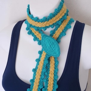 May include: A handmade crochet scarf in teal and tan. The scarf features a leaf-shaped accent and a scalloped edge. The scarf is displayed on a mannequin wearing a navy blue tank top.