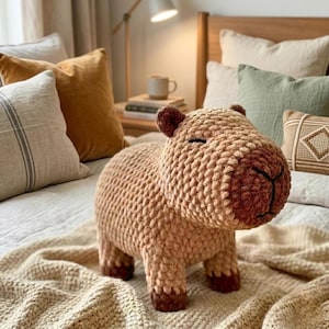 May include: A crocheted capybara soft toy in tan and brown hues. The cuddly animal has a rounded body, small ears, and closed eyes, placed on a bed with pillows and a blanket. The capybara is a large rodent native to South America.