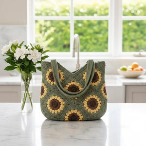 May include: A handmade green crochet tote bag with sunflower designs. The bag features brown and yellow sunflower appliques on a green background. The tote has two handles and is sitting on a white surface.