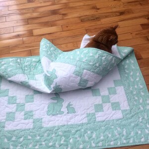 May include: A green and white quilt with a checkered pattern and a design of bunnies. A cat is curled up inside the quilt.