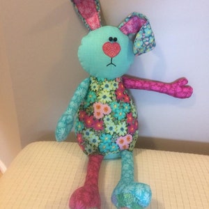 K&ouml;nnte beinhalten: Ein handgemachtes Pl&uuml;schtier-H&auml;schen mit einem blauen und rosa floralen Muster. Das H&auml;schen hat lange Arme und Beine und einen gro&szlig;en Kopf mit einer rosa Herznase und schwarzen Augen.