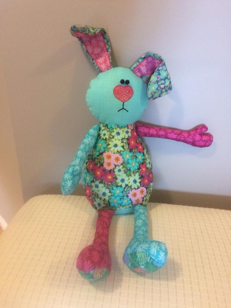 K&ouml;nnte beinhalten: Ein handgemachtes Pl&uuml;schtier-H&auml;schen mit einem blauen und rosa floralen Muster. Das H&auml;schen hat lange Arme und Beine und einen gro&szlig;en Kopf mit einer rosa Herznase und schwarzen Augen.