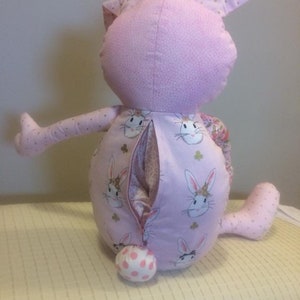 K&ouml;nnte beinhalten: Ein rosa Pl&uuml;sch-Hasenspielzeug mit einem Rei&szlig;verschluss auf der R&uuml;ckseite. Der Hase hat einen wei&szlig;en Tupfen-Schwanz und ist mit einem rosa Stoff mit einem Muster aus wei&szlig;en Hasen bedeckt.