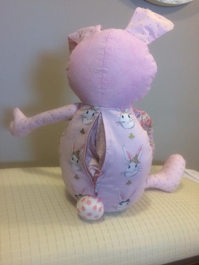 K&ouml;nnte beinhalten: Ein rosa Pl&uuml;sch-Hasenspielzeug mit einem Rei&szlig;verschluss auf der R&uuml;ckseite. Der Hase hat einen wei&szlig;en Tupfen-Schwanz und ist mit einem rosa Stoff mit einem Muster aus wei&szlig;en Hasen bedeckt.