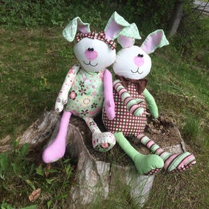 K&ouml;nnte beinhalten: Zwei handgefertigte Pl&uuml;schhasen sitzen auf einem Baumstumpf. Die Hasen tragen rosa und gr&uuml;n gestreifte Kleidung mit Blumen- und Tupfenmustern.