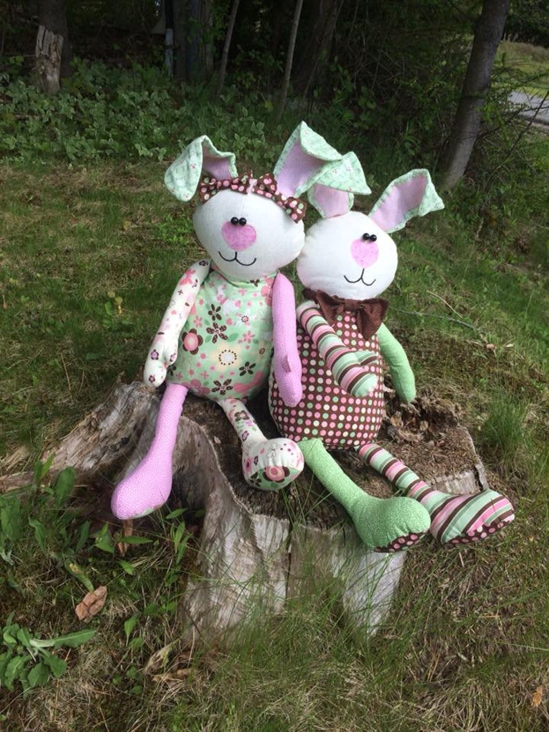 K&ouml;nnte beinhalten: Zwei handgefertigte Pl&uuml;schhasen sitzen auf einem Baumstumpf. Die Hasen tragen rosa und gr&uuml;n gestreifte Kleidung mit Blumen- und Tupfenmustern.