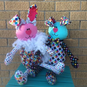 K&ouml;nnte beinhalten: Zwei handgefertigte Pl&uuml;schtiere, ein rosa Kaninchen und ein blauer Hund, sitzen auf einer t&uuml;rkisfarbenen Bank. Beide Spielzeuge sind in bunten Patchwork-Stoff mit geometrischen Mustern und Punkten gekleidet. Das Kaninchen hat eine wei&szlig;e Federboa um den Hals und eine rosa Schleife auf dem Kopf.