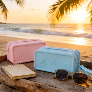 May include: A light pink and a light blue rectangular cosmetic bag with a zipper closure, a small notebook, and sunglasses on a wooden tray. The bags have a name and a seashell design. The background shows a beach and ocean.
