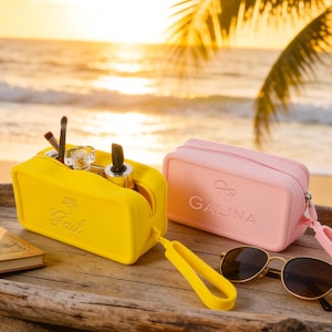 May include: A yellow makeup bag with the name "Fail" embossed on the front, filled with makeup brushes and products. A pink makeup bag with "GALINA" printed on it sits next to it. Sunglasses and a notebook are also on the wooden surface.